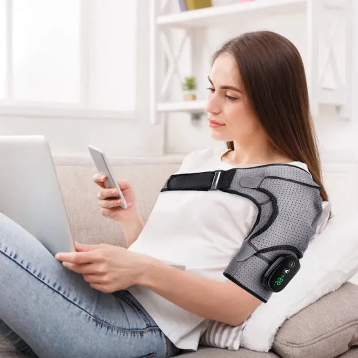 Woman relaxing at home using the RelaxRiser™ HeatFlex Shoulder Wrap while working on a laptop and phone, showcasing its hands-free and comfortable design for everyday relief