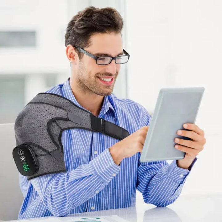 Man using the RelaxRiser™ HeatFlex Shoulder Wrap while working on a tablet, demonstrating hands-free use and ergonomic comfort for shoulder pain relief.